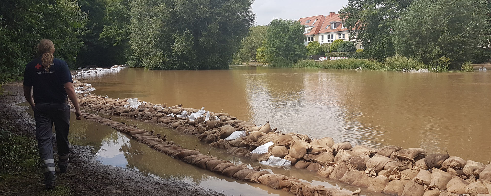 Blick auf die Innerste bei Hochwasser. Das Flussufer ist mit Sandsäcken gesichert. Links ist eine DRK-Einsatzkraft von hinten zu sehen.
