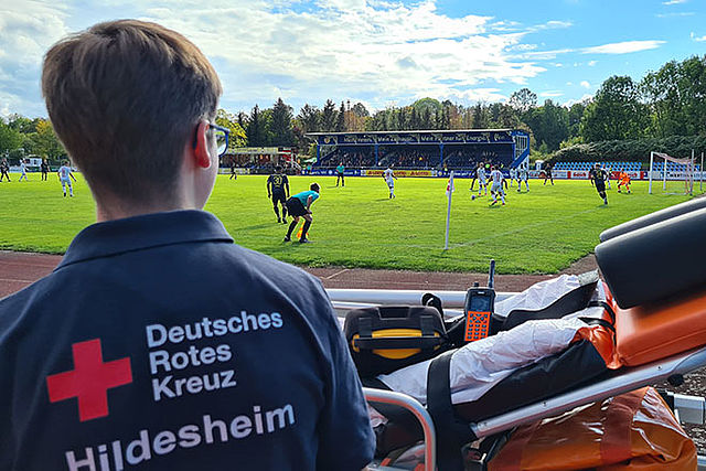 Sanitätsdienst bei Fußballspiel. Im Vordergrund sind ein Sanitäter von hinten mit großem DRK-Logo auf dem Rücken sowie eine Trage zu sehen. Hintergrund bildet das Spielfeld mit Spielern.