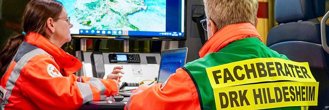 Einsatzkraft mit Fachberater-Weste an der Seitentür eines ELW. Darin sitzt weitere Einsatzkraft, dahinter sind Funkgeräte und ein Monitor zu sehen.