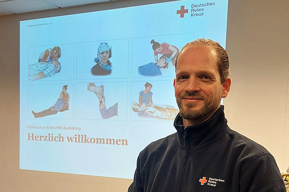 Der Ausbilder steht im Unterrichtsraum und blickt freundlich in die Kamera. Hinter ihm ist eine Folie aus der Ausbildung an die Wand projiziert. Herzlich willkommen steht darauf.