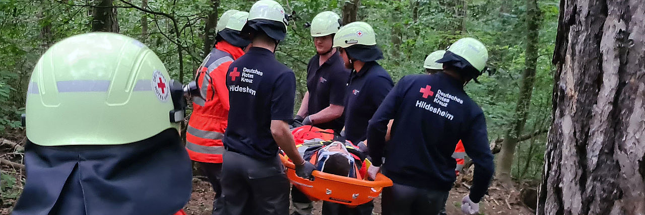 Sanitäter tragen einen Verletzten in einer Schleifkorbtrage durch den Wald. Im Vordergrund ist eine Einsatzkraft mit leuchtroter Jacke mit rotem Kreuz von hinten abgebildet.