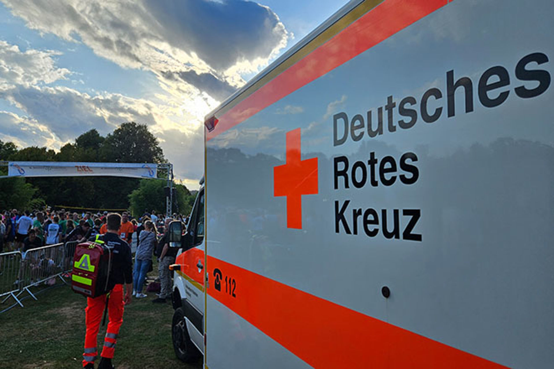Sanitätsdienst bei Laufsport-Veranstaltung. Ein Sanitäter und ein RTW sind von hinten abgebildet. Im Hintergrund Läufer.