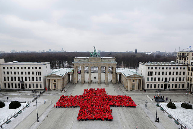 Riesige rotes Kreuz bestehend aus Menschen vor dem Brandenburger Tor in Berlin, schräg von oben aufgenommen.
