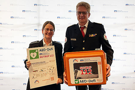 Ute und Heiko Stillahn bei der Übergabe des Defis. Beide stehen im Dienstanzug mit Plakat bzw. Defi vor einer Werbewand der Volksbank.