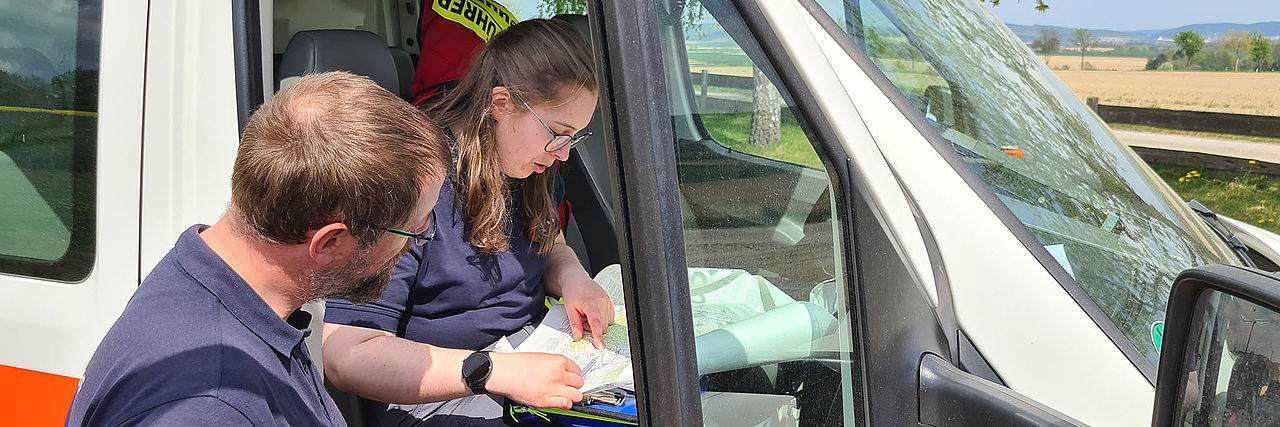 Nahaufnahme von zwei Einsatzkräften an bzw. in einem Fahrzeug. Beide sind nach vorn gebeugt und arbeiten an einer Karte.