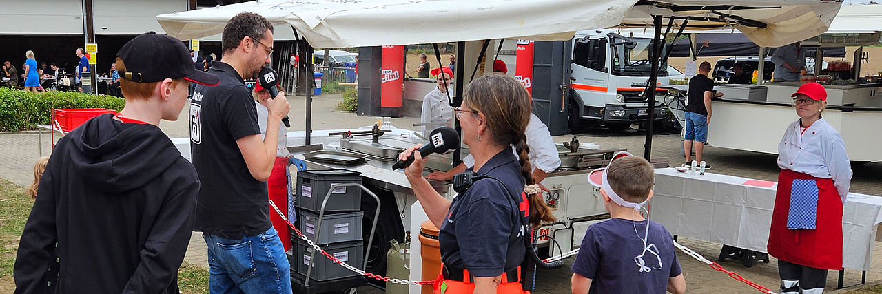 FFN-Moderator im Interview mit einer Einsatzkraft vor der laufenden Feldküche.