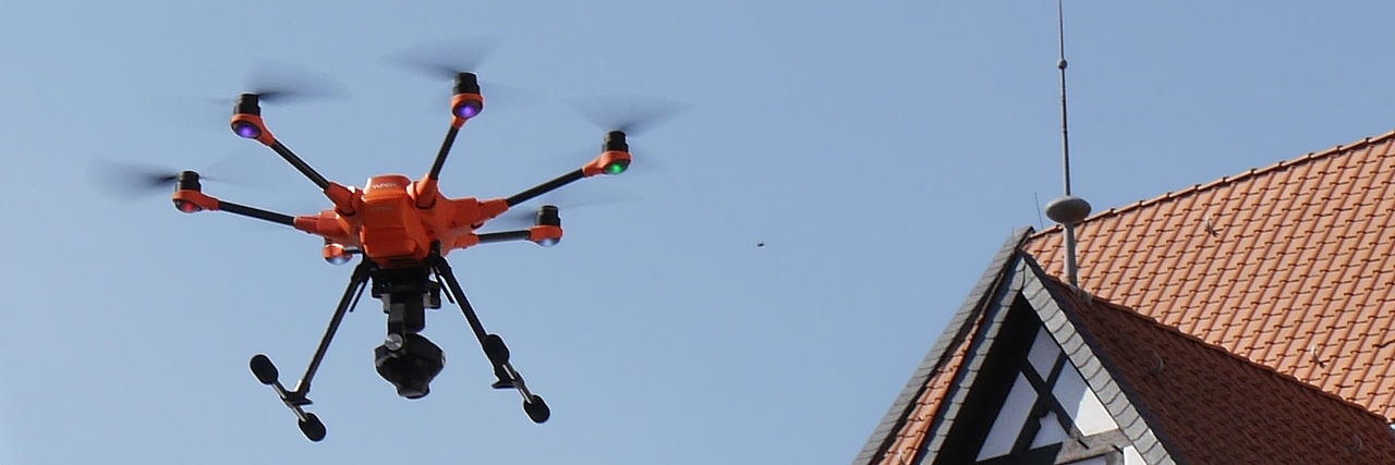 Orangefarbene Drohne vor blauem Himmel. Auf einer Seite ist der Giebel eines Hauses zu sehen.