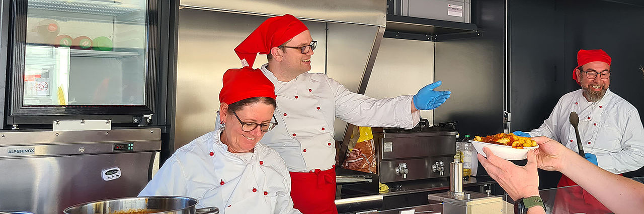 Drei DRK-Einsatzkräfte mit weißen Kochjacken und roten Kopftüchern arbeiten im Food-Truck.