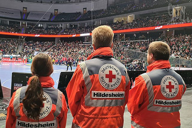 Drei Sanitäter stehen vor einer Halle, in der ein Handballspiel stattfindet. Sie sind groß von hinten zusehen. Spielfeld und Ränge mit Publikum sind zu erkennen.