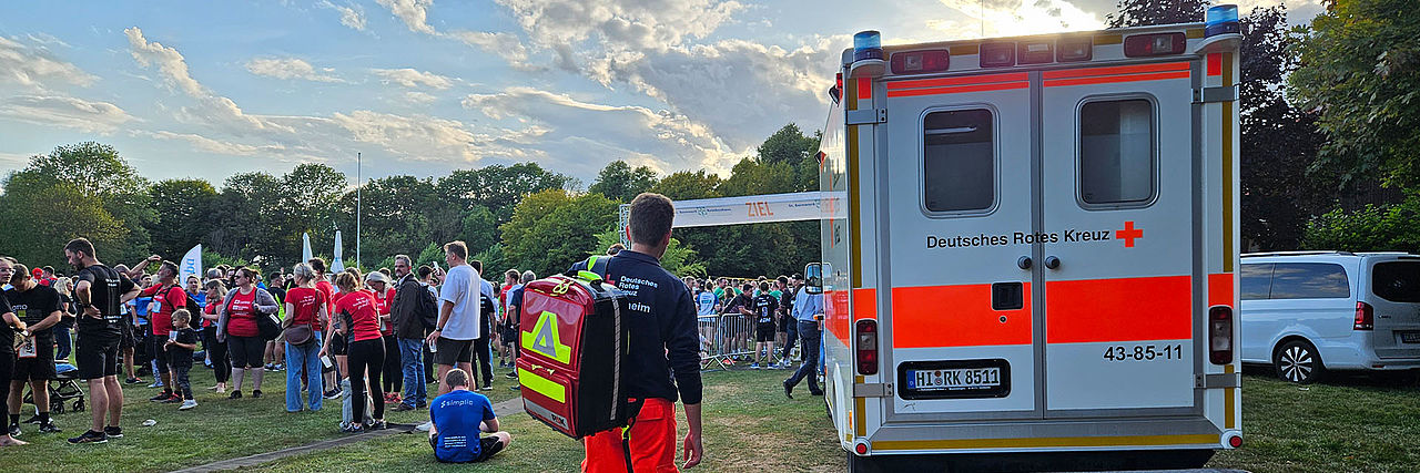 Sanitätsdienst bei Laufsport-Veranstaltung. Ein Sanitäter und ein RTW sind von hinten abgebildet. Im Hintergrund Läufer.