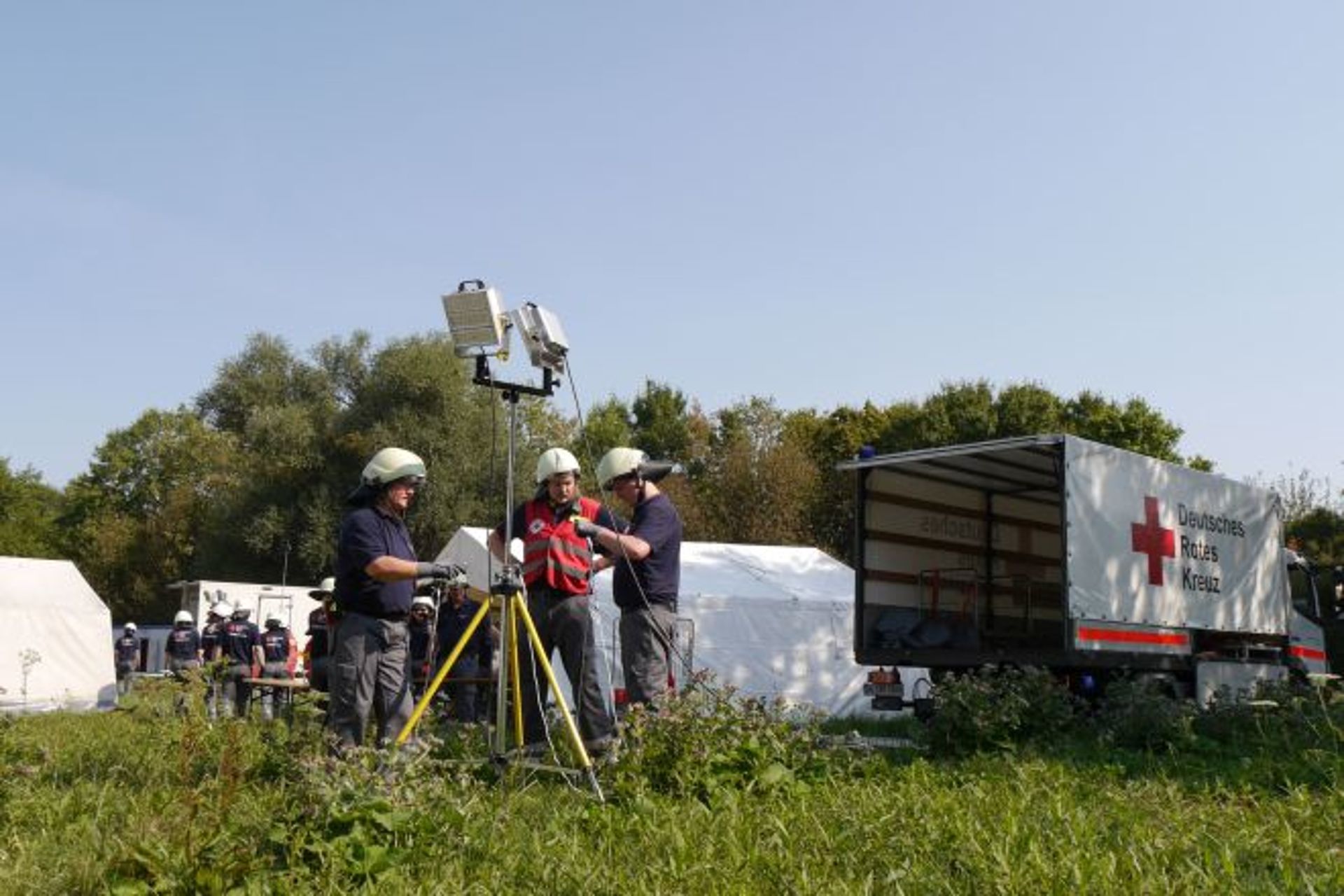 Einsatzkräfte bauen ein Stativ mit Scheinwerfer auf einer Wiese auf.