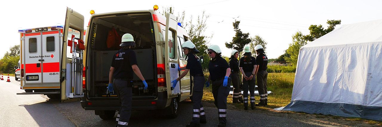 KTW steht mit geöffneten Hecktüren am Behandlungsplatz. Mehrere Einsatzkräfte befinden sich zwischen Fahrzeug und Behandlungszelt.
