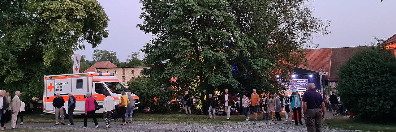 Sanitätsdienst bei einem klassischen Konzert abends unter freiem Himmel. Rechts ist der DRK-RTW zusehen, viele Menschen laufen umher.